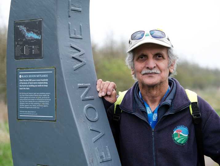 Kelly Sagar, wearing outdoor walking gear, a hat and sunglasses, stands by an information post on the Black Devon Wetlands.