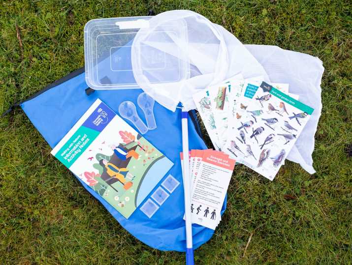 A collection of Walking with Nature and Strength and Balance resources include a bag, booklets, leaflets, net with handle and a plastic tub with a grassy ground as a backdrop.