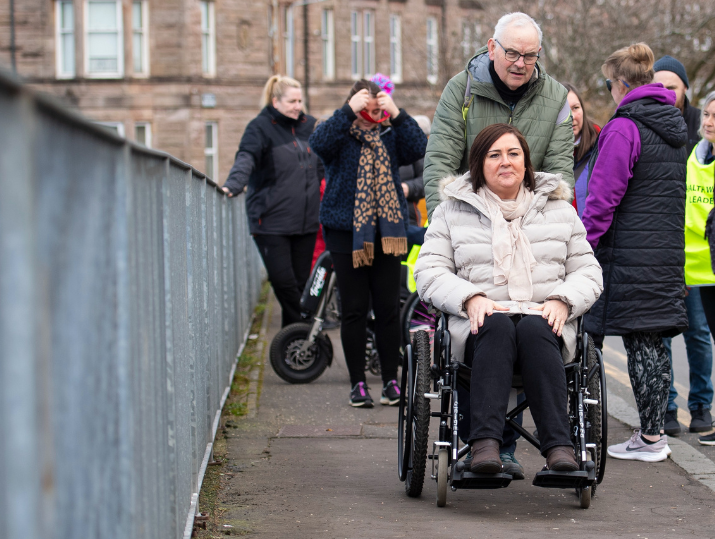 A group of participants on a Health Walk wearing blindfolds, demo glasses and using wheelchairs smile as they walk along a street.