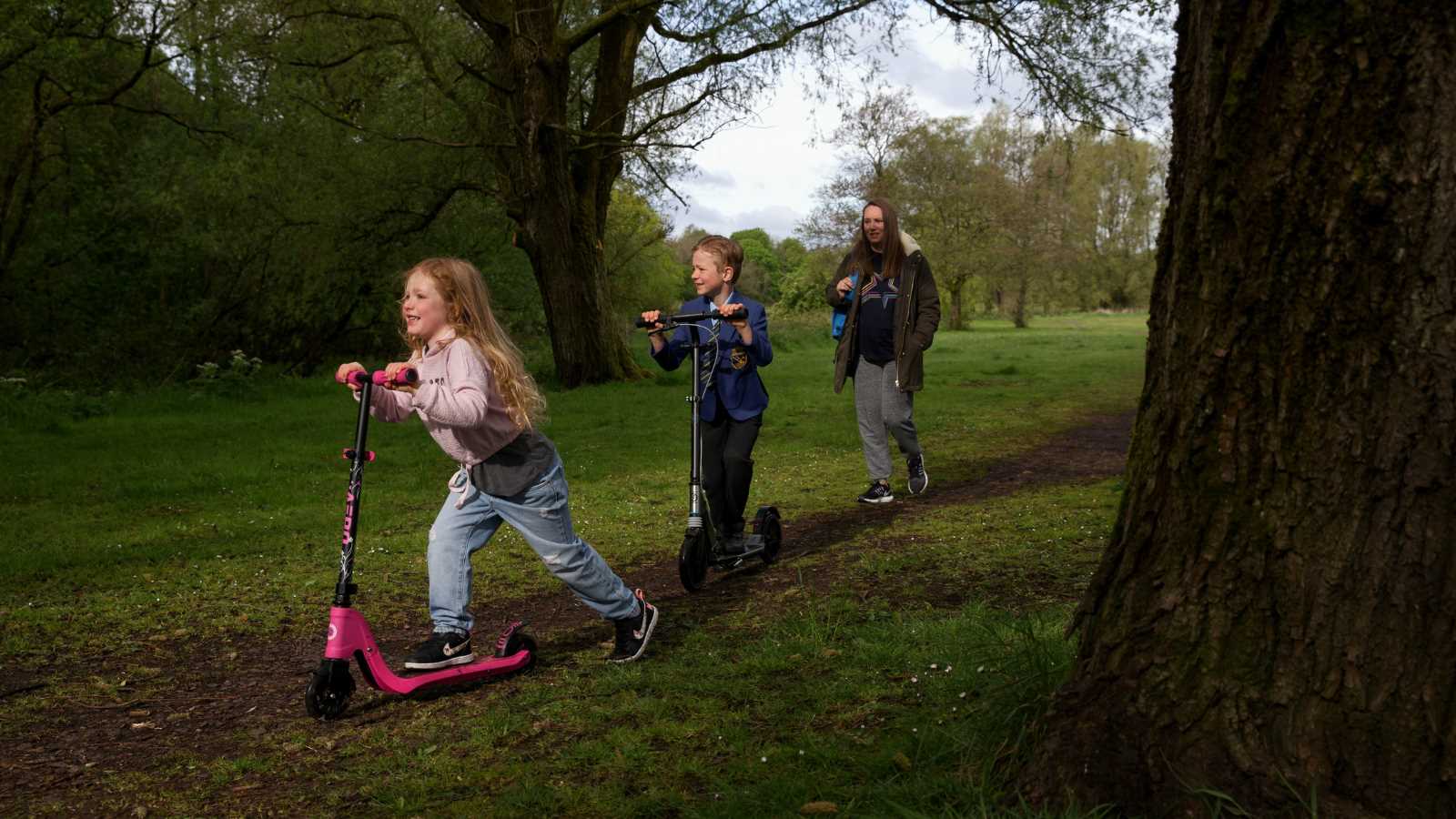 Two young children smile as they scoot along a path, followed by a parent or guardian.